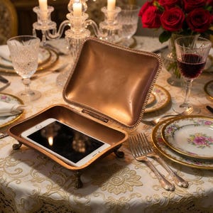 May include: A copper-toned, hinged box containing a smartphone, displayed on a table set with fine china, silverware, and red roses. The box is open, showing the phone's screen. The table is covered with a white tablecloth.