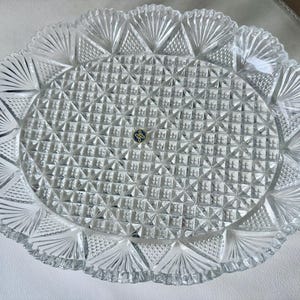 May include: Clear, oval-shaped glass serving platter with a textured, geometric pattern. The rim features a scalloped edge with fan-like designs. A small, circular emblem is visible in the center. This decorative piece is suitable for serving food.