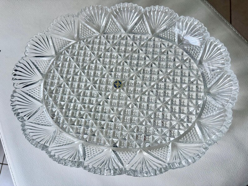May include: Clear, oval-shaped glass serving platter with a textured, geometric pattern. The rim features a scalloped edge with fan-like designs. A small, circular emblem is visible in the center. This decorative piece is suitable for serving food.