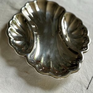 Pode incluir: Um prato de servir em metal prateado polido com um design festonado em forma de concha, apresentando tr&ecirc;s compartimentos curvos distintos. A superf&iacute;cie &eacute; altamente reflexiva, exibindo bordas suaves e arredondadas e uma forma sim&eacute;trica e org&acirc;nica.