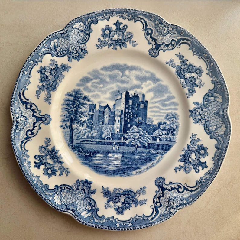 May include: A round ceramic plate with a scalloped edge featuring a blue and white transferware pattern. The center depicts a castle by a lake with the text Blarney Castle. The border is decorated with intricate floral and scrollwork motifs.