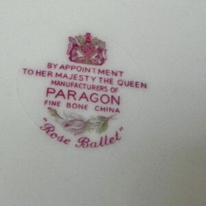 May include: A white ceramic surface featuring a pink printed maker's mark for Paragon Fine Bone China. The text reads BY APPOINTMENT TO HER MAJESTY THE QUEEN MANUFACTURERS OF PARAGON FINE BONE CHINA with a rose illustration and the text Rose Ballet.