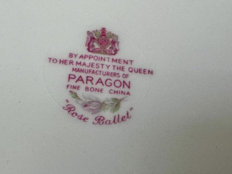 May include: A white ceramic surface featuring a pink printed maker's mark for Paragon Fine Bone China. The text reads BY APPOINTMENT TO HER MAJESTY THE QUEEN MANUFACTURERS OF PARAGON FINE BONE CHINA with a rose illustration and the text Rose Ballet.