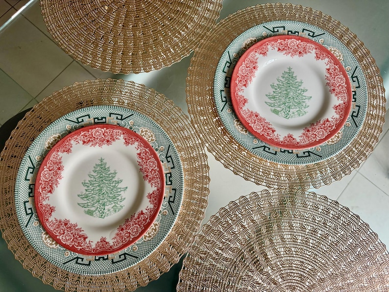May include: Overhead shot of a table setting with four place settings. Each setting includes a gold-colored placemat, a green and white patterned plate, and a smaller plate with a red floral border and a green Christmas tree design.