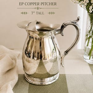 May include: A silver-colored metal pitcher with a curved handle and a wide spout. The text VINTAGE PRIMROSE PLATE EP COPPER PITCHER 7" TALL is visible above the object. The pitcher has a polished, reflective surface and a circular base.