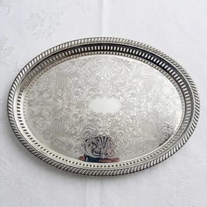 May include: An oval silver-toned serving tray featuring an intricate engraved floral and scrollwork pattern across the center. The tray has a raised, rope-textured border and a pierced gallery rim with a series of vertical rectangular slots.