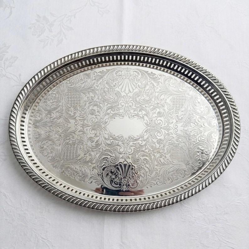 May include: An oval silver-toned serving tray featuring an intricate engraved floral and scrollwork pattern across the center. The tray has a raised, rope-textured border and a pierced gallery rim with a series of vertical rectangular slots.