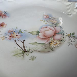 May include: White ceramic serving dish with a floral design. The hand-painted flowers include pink peonies, peach blossoms, and yellow daisies, with green leaves and brown stems. The dish has a scalloped edge and a handle.