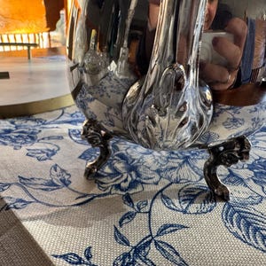 May include: A polished silver teapot featuring a curved handle with ornate leaf-like detailing at the base. The teapot stands on three small, decorative feet and has a highly reflective surface that shows a distorted reflection of the surroundings.