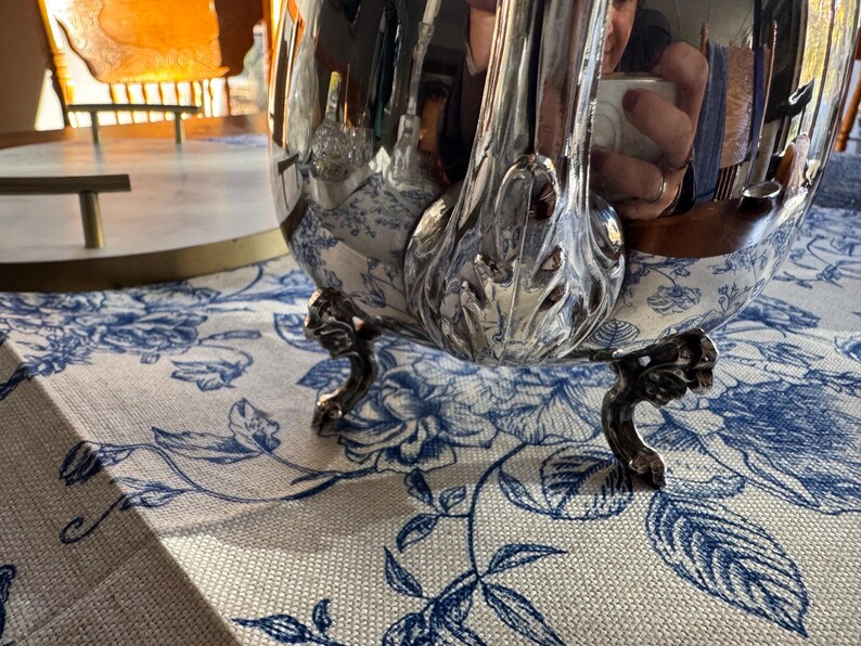 May include: A polished silver teapot featuring a curved handle with ornate leaf-like detailing at the base. The teapot stands on three small, decorative feet and has a highly reflective surface that shows a distorted reflection of the surroundings.
