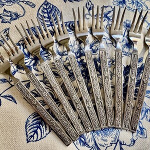 May include: A set of eight silver-toned metal forks arranged in a fan pattern. Each fork features a long, slender handle with an embossed, intricate geometric and floral pattern running down the centre, ending in a four-tined head.
