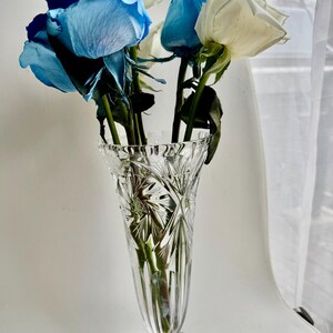 May include: A clear, cut-glass vase with a flared, scalloped rim and a starburst pattern on the base. The vase contains a bouquet of several long-stemmed roses, featuring a mix of vibrant blue and creamy white petals with green foliage.