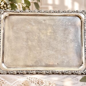 Puede incluir: Una bandeja de servicio rectangular de tono plateado con una superficie central lisa y pulida, y un borde elevado decorado con intrincados detalles florales y de volutas a lo largo de los cuatro lados del marco.