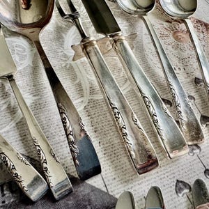 以下が含まれることがあります： パターン化された表面に配置された、スプーン、ナイフ、フォークを含むアンティークシルバーカトラリーの品揃え。銀食器は、ハンドルに装飾的な花のディテールが施されています。コレクションには、バターナイフとサービングスプーンが含まれています。
