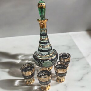 May include: A set of four clear glass shot glasses with gold trim and a matching green and gold glass decanter. The glasses and decanter have a black and white striped design with floral accents.