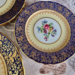 May include: A collection of antique porcelain plates. The plates feature a cream-colored center with a floral bouquet and a navy blue border with gold leaf detailing. The rims are scalloped and trimmed with gold.