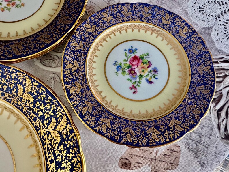 May include: A collection of antique porcelain plates. The plates feature a cream-colored center with a floral bouquet and a navy blue border with gold leaf detailing. The rims are scalloped and trimmed with gold.
