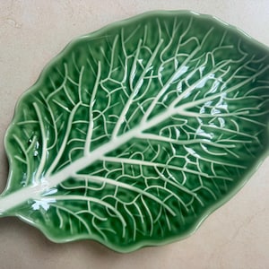 Puede incluir: Un plato de cer&aacute;mica verde brillante con forma de hoja de col. El plato presenta una nervadura central blanca prominente y elevada, con nervaduras blancas m&aacute;s peque&ntilde;as y ramificadas que se extienden por la superficie texturizada y festoneada.