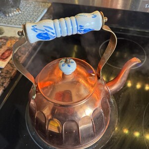 May include: A copper tea kettle with a fluted base and a curved spout. The kettle features a white ceramic handle and a matching lid knob, both decorated with blue floral patterns. The metal handle frame is brass-toned and arched over the top.