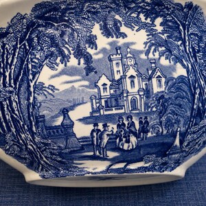 Op de afbeelding: Een witte keramische terrine met een gedetailleerd blauw transferpatroon. Het ontwerp toont een groot kasteel omringd door bomen, met verschillende figuren op de voorgrond. De sc&egrave;ne is omlijst door dicht gebladerte en architecturale elementen.