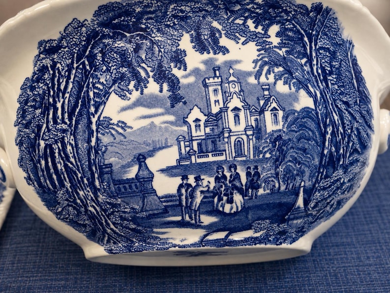 Op de afbeelding: Een witte keramische terrine met een gedetailleerd blauw transferpatroon. Het ontwerp toont een groot kasteel omringd door bomen, met verschillende figuren op de voorgrond. De sc&egrave;ne is omlijst door dicht gebladerte en architecturale elementen.