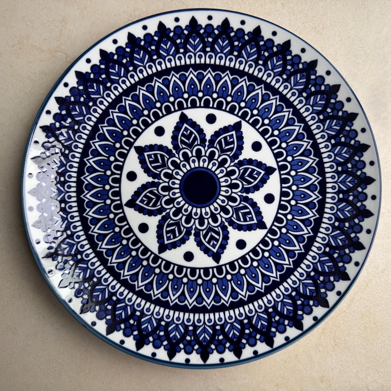 May include: A round ceramic plate featuring a detailed, symmetrical mandala pattern in shades of deep blue and white. The design consists of concentric rings of intricate leaf-like shapes, dots, and geometric lines radiating from a solid dark blue center.