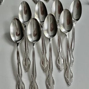 May include: A collection of silver spoons with ornate, floral-like detailing on the handles. The spoons are arranged in a neat, organized display, reflecting light. The spoons are likely part of a vintage silverware set.