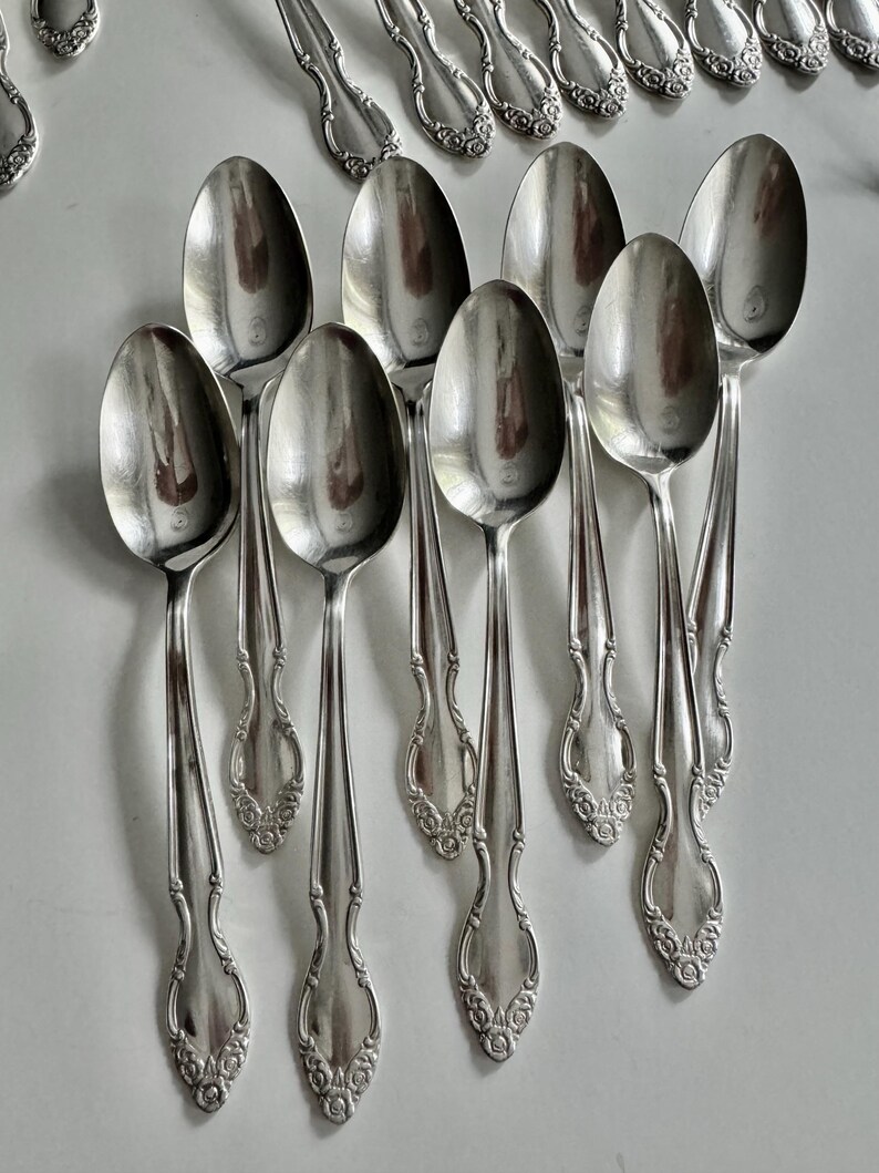 May include: A collection of silver spoons with ornate, floral-like detailing on the handles. The spoons are arranged in a neat, organized display, reflecting light. The spoons are likely part of a vintage silverware set.