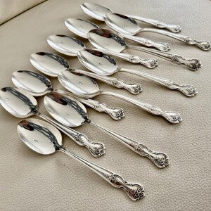 May include: A collection of silver spoons arranged in a diagonal pattern. The spoons have a reflective surface and ornate floral detailing on the handles. The spoons are set against a neutral background.