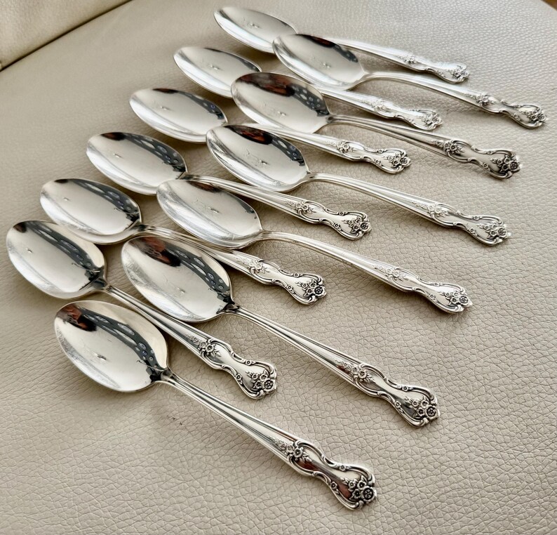 May include: A collection of silver spoons arranged in a diagonal pattern. The spoons have a reflective surface and ornate floral detailing on the handles. The spoons are set against a neutral background.