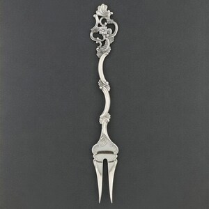 K&ouml;nnte beinhalten: Eine silberne Gabel mit einem verzierten, floralen Design am Griff. Der Griff ist anmutig geschwungen und f&uuml;hrt zu zwei Zinken. Die Gabel ist wahrscheinlich f&uuml;r das Servieren oder einen besonderen Anlass gedacht. Der Hintergrund ist einfarbig dunkelgrau.