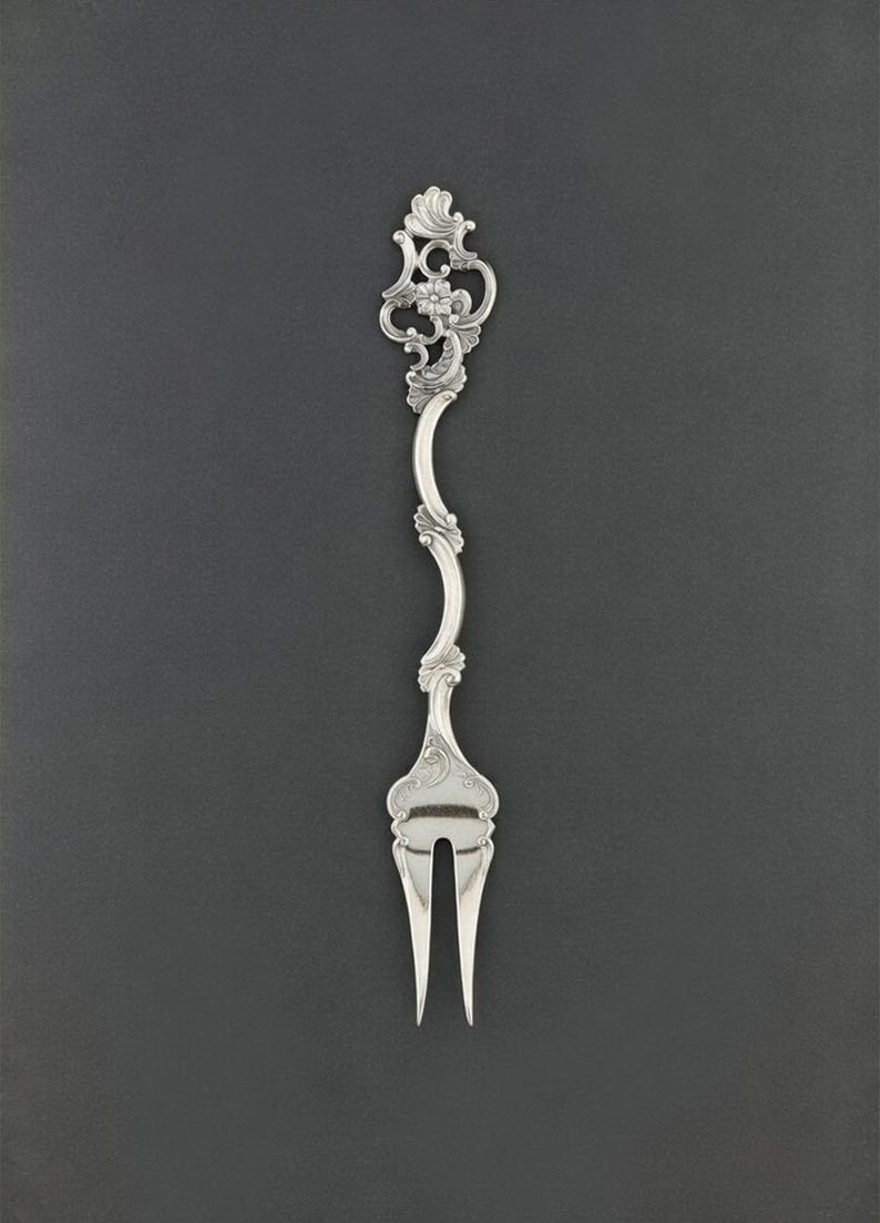K&ouml;nnte beinhalten: Eine silberne Gabel mit einem verzierten, floralen Design am Griff. Der Griff ist anmutig geschwungen und f&uuml;hrt zu zwei Zinken. Die Gabel ist wahrscheinlich f&uuml;r das Servieren oder einen besonderen Anlass gedacht. Der Hintergrund ist einfarbig dunkelgrau.