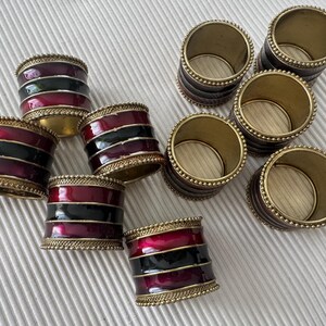 May include: A collection of napkin rings with a burgundy, black, and gold design. The rings have a metallic gold trim with a beaded edge. The napkin rings are cylindrical and appear to be made of metal. The rings are suitable for a dining table.