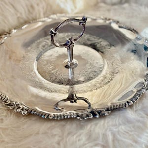 May include: A silver-coloured, two-tiered serving tray with an ornate handle. The tray has a decorative, scalloped edge and a detailed floral pattern. The handle is curved, adding to the vintage aesthetic. The tray is likely made of metal.
