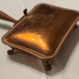 May include: A vintage copper-toned crumb tray with a wooden handle and ornate brass feet. The rectangular tray has a decorative edge and a small brass clasp. The tray is designed to collect crumbs.