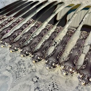 May include: A collection of ornate silver knives arranged in rows. The knives have a shiny, reflective blade and a detailed, decorative handle. The handles feature a floral design. The knives are displayed on a white lace tablecloth.