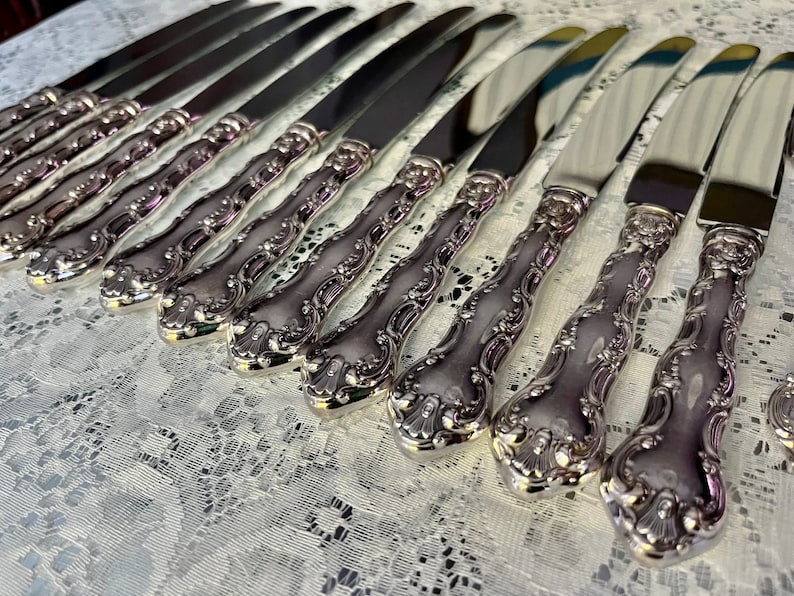 May include: A collection of ornate silver knives arranged in rows. The knives have a shiny, reflective blade and a detailed, decorative handle. The handles feature a floral design. The knives are displayed on a white lace tablecloth.