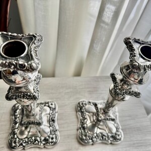 May include: A pair of silver-colored metal candlesticks featuring ornate, embossed floral and scrollwork detailing on the square bases and the top candle holders. The stems are tapered with decorative tiered sections and a polished finish.