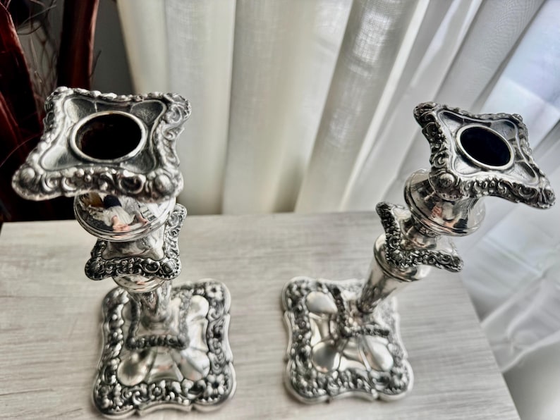 May include: A pair of silver-colored metal candlesticks featuring ornate, embossed floral and scrollwork detailing on the square bases and the top candle holders. The stems are tapered with decorative tiered sections and a polished finish.