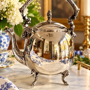 May include: A polished silver tea kettle featuring an ornate, curved handle and a matching spout. The kettle has a rounded body with a reflective surface, a decorative lid finial, and stands on three intricate, scrolled metal feet.