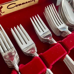 May include: A set of silver silverware, including forks and spoons, arranged in a red velvet-lined box. The forks and spoons are neatly organized, with a decorative pattern on the handles. The box has a gold label that reads "Community The Finest Silverplate."