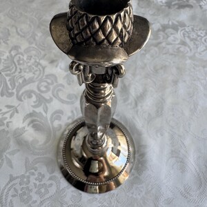 May include: A silver-toned metal candlestick holder featuring a top section shaped like a textured hat with a diamond-patterned crown. The stem has a smooth, tapered design leading down to a circular base with a beaded edge detail along the rim.