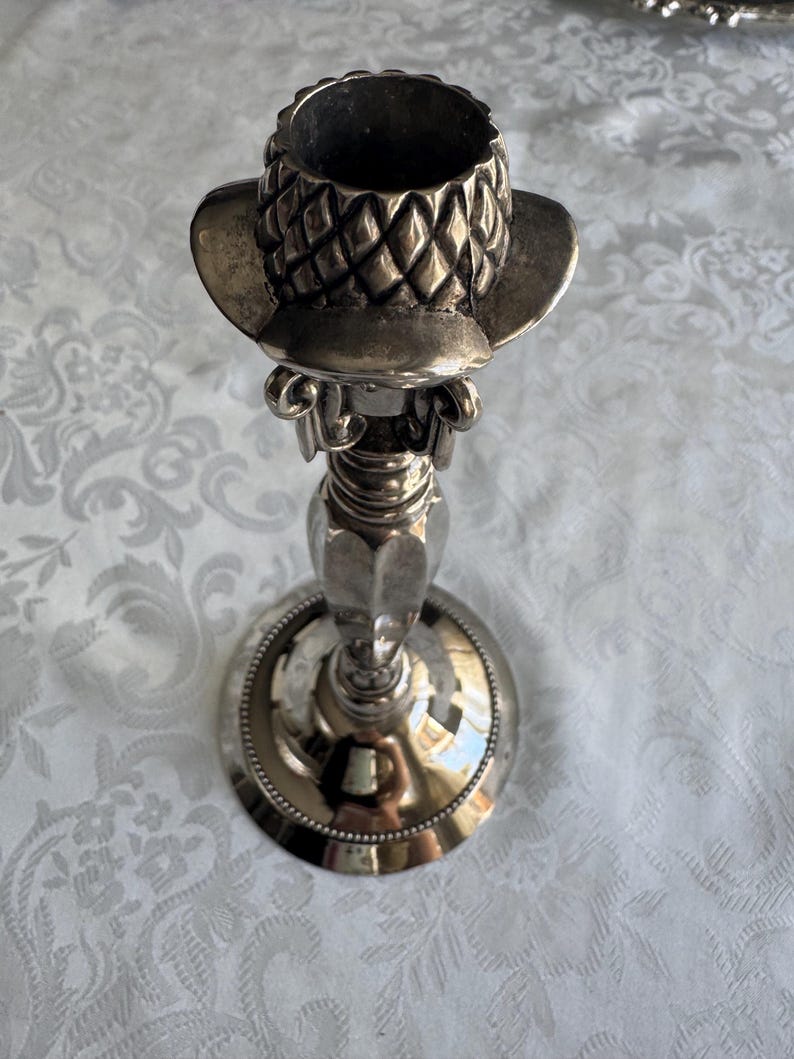 May include: A silver-toned metal candlestick holder featuring a top section shaped like a textured hat with a diamond-patterned crown. The stem has a smooth, tapered design leading down to a circular base with a beaded edge detail along the rim.