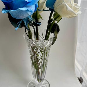 May include: A clear, cut-glass vase with a flared rim and a starburst pattern on the base, containing a bouquet of vibrant blue and white roses with green stems and leaves. The glass features intricate geometric facets that catch the light.