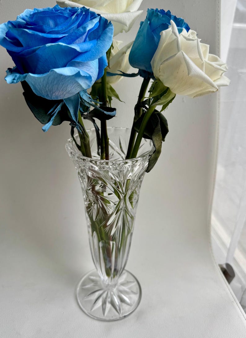 May include: A clear, cut-glass vase with a flared rim and a starburst pattern on the base, containing a bouquet of vibrant blue and white roses with green stems and leaves. The glass features intricate geometric facets that catch the light.