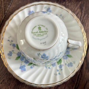 May include: A white teacup and saucer set with a gold trim and a floral pattern of blue flowers and green leaves. The teacup is upside down on the saucer. The bottom of the teacup has the text "Royal Adderley" and "Fine Bone China England".