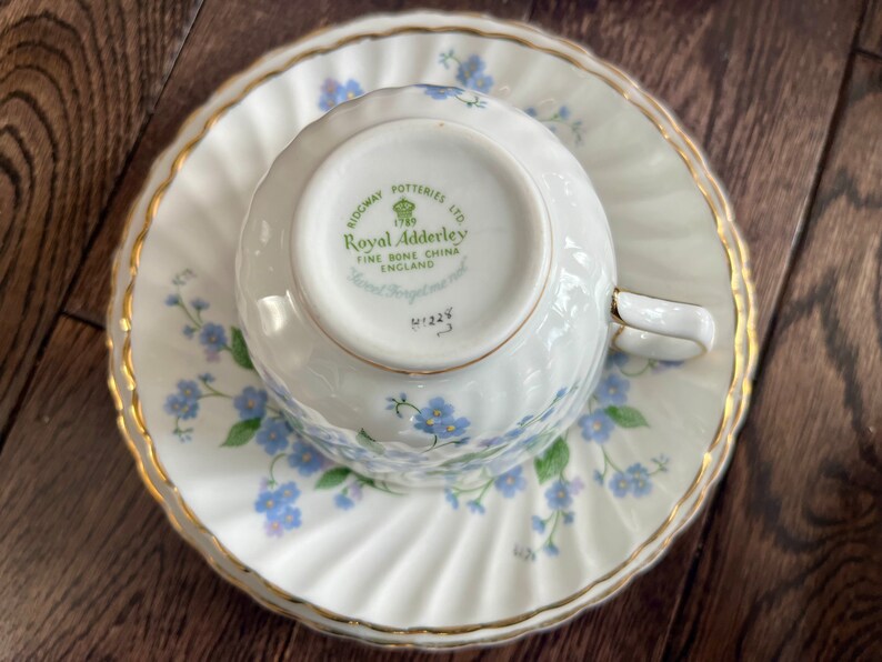May include: A white teacup and saucer set with a gold trim and a floral pattern of blue flowers and green leaves. The teacup is upside down on the saucer. The bottom of the teacup has the text "Royal Adderley" and "Fine Bone China England".