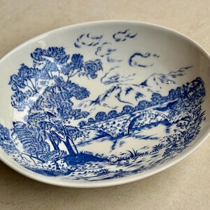 May include: Oval-shaped, white ceramic bowl with a blue landscape design. The design features trees, a bridge, mountains, and clouds. The bowl has a smooth, glossy finish and is likely for serving food.