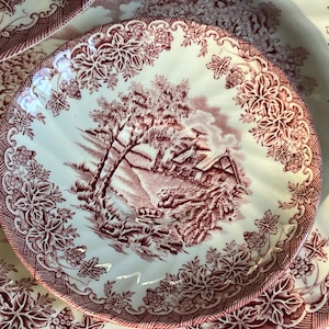 May include: A set of three vintage ceramic plates with a red and white floral pattern. The plates feature a cottage scene with trees and a stream.