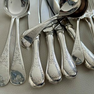 May include: An assortment of antique silverware, including spoons, knives, and a ladle, all with a shiny, reflective surface. Each piece is adorned with an intricate floral pattern, adding a touch of elegance. The silverware is arranged on a neutral-toned surface.